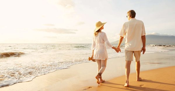 A man and woman stroll hand in hand along the beach, with gentle waves lapping at the shore in the background.