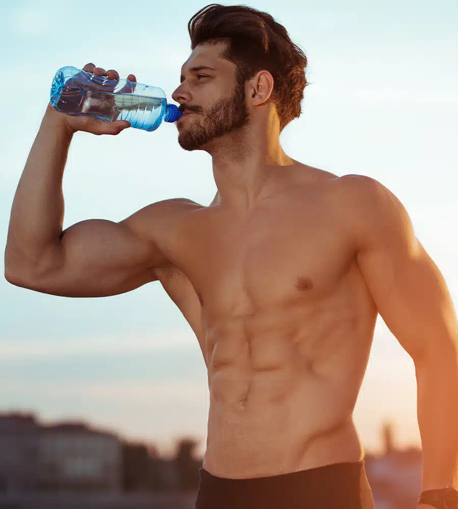 A shirtless man drinks water from a bottle, standing outdoors in a casual setting.