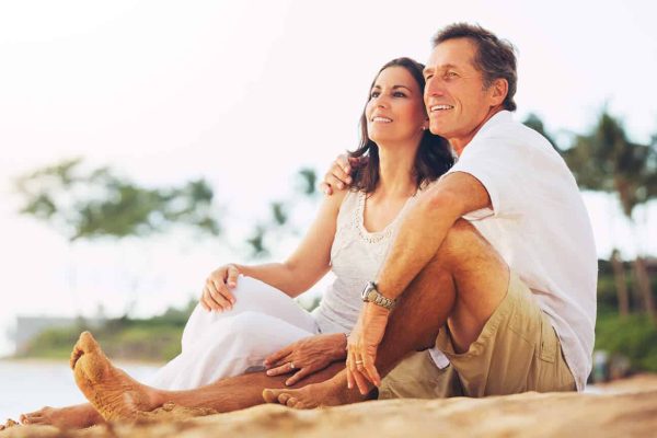 A couple sits on the sandy beach, surrounded by the sea and a clear blue sky.